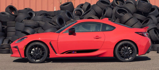 A bright red sports coupe car shown in side profile, parked outdoors in front of a large pile of black tires.