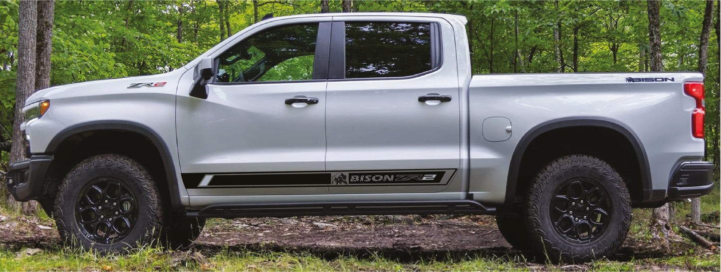 A white pickup truck with black wheels and side decals is parked outdoors on a grassy path surrounded by green trees. The side decal reads 'BISON'.