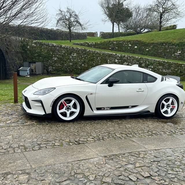 White sports car parked on a stone path with a stone wall and trees in the background