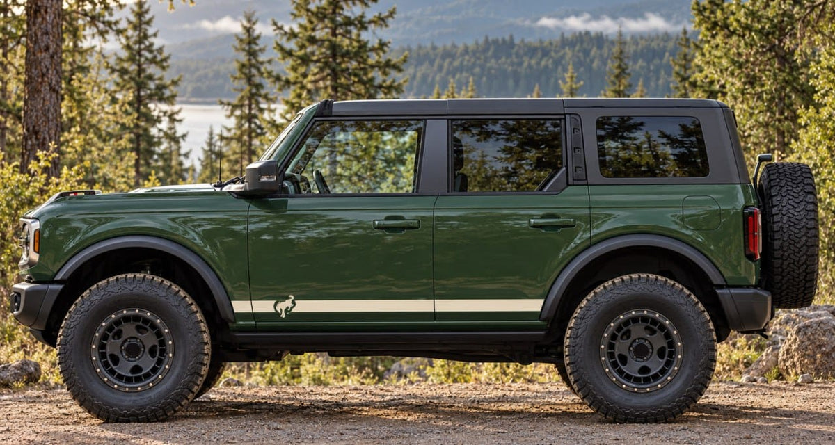 A dark green Ford Bronco SUV with off-road tires and a spare tire mounted on the rear door, parked outdoors on a gravel path with trees and mountains in the background.