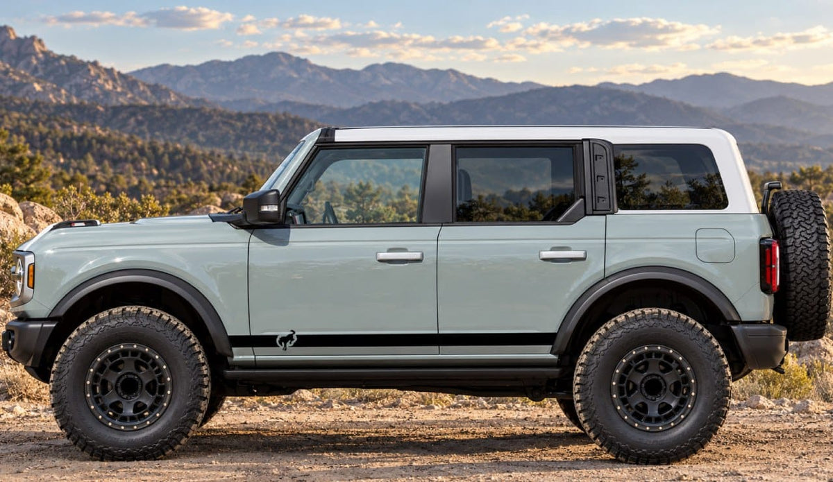 A light sage green Ford Bronco with a white roof stands in profile on a dirt road with mountains in the background.