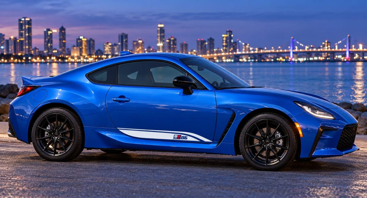 A bright blue sports coupe parked outdoors at dusk with a city skyline and bridge reflected in the water behind it. The car has black wheels, a white side stripe, and a small M20 badge near the rear door.