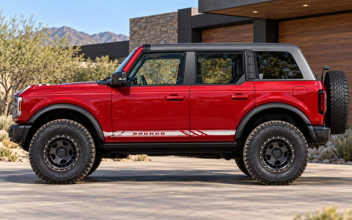 A red Ford Bronco SUV with black off-road tires and a black soft top, parked outdoors in front of a modern house with mountains in the background.