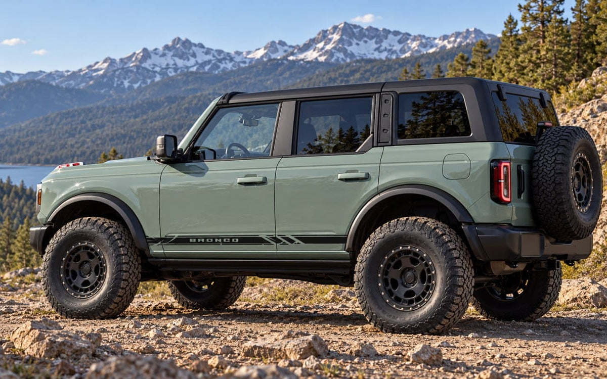 A sage green Ford Bronco with black trim and off-road tires parked on a rocky mountain path with snow-capped peaks and a lake in the background.
